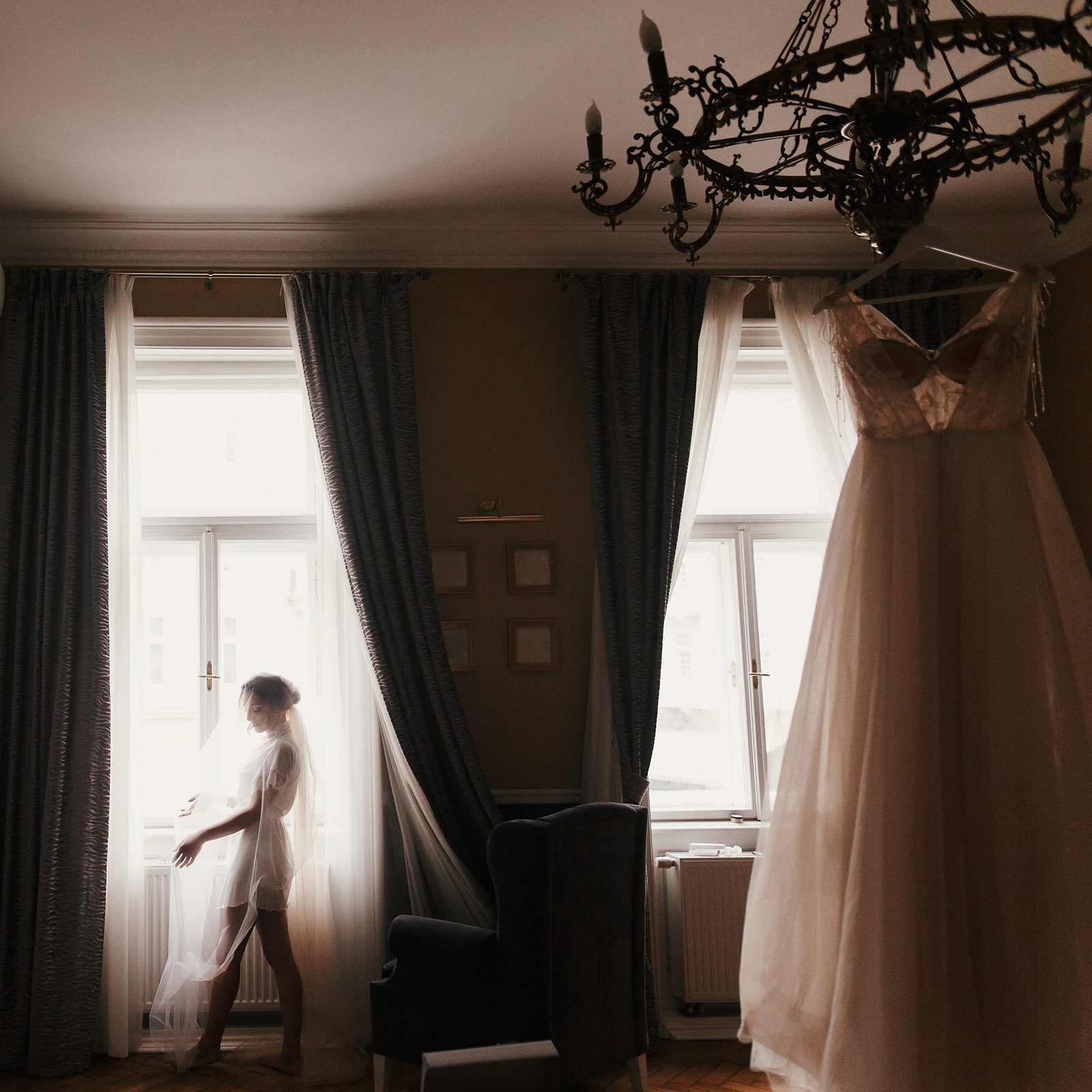 Gorgeous bride silhouette standing at window light, looking at stylish wedding dress, hanging on chandelier, in the morning. Bride and her beautiful wedding gown. Woman getting ready
